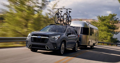 A Subaru Ascent SUV pulls a silver camper with two bicycles on its roof rack