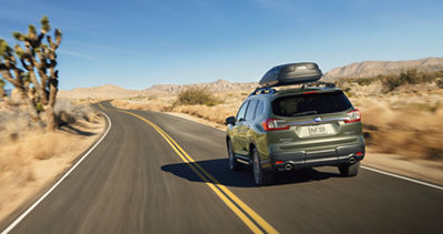 A Subaru Ascent large SUV with a rooftop cargo carrier drives on a desert highway.