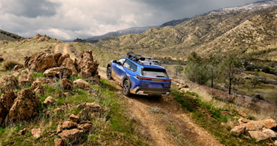 A static image of a blue 2026 Subaru Trailseeker driving on a narrow dirt trail that winds through rocky, grassy hills.