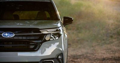 The front of a 2026 Subaru Forester AWD SUV in silver, half in view, highlighting the Forester’s headlights, grille, Subaru logo, and windshield.