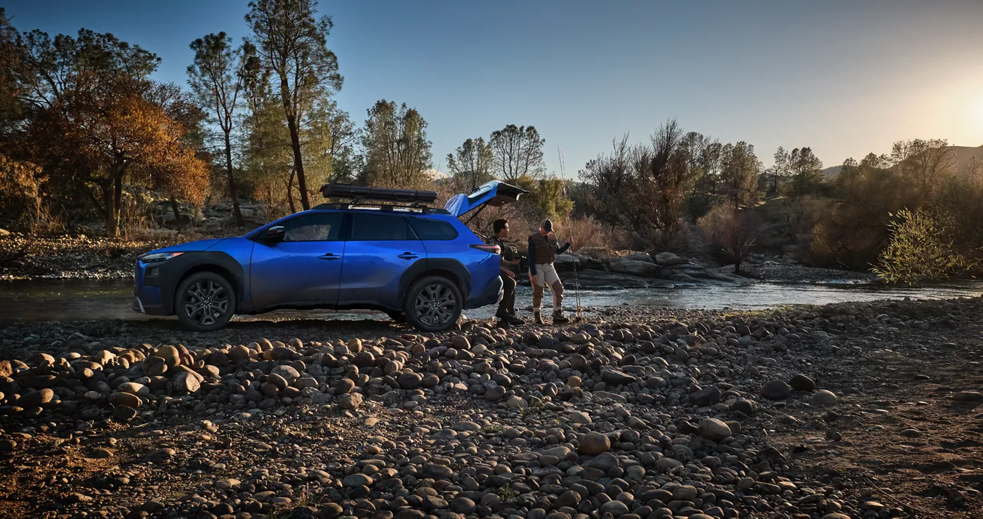 A blue Subaru Trailseeker parked at a river as two people unload gear