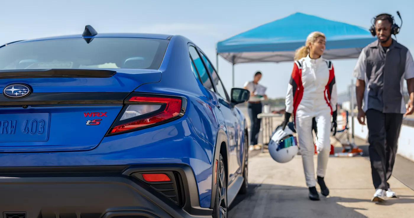 A blue Subaru WRX in a paddock as two people in racing gear walk past