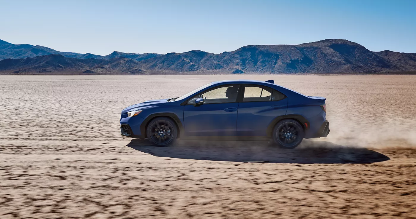 A blue Subaru WRX in profile kicking up dust on a dry lake bed