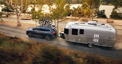 A Subaru Ascent family SUV tows a large silver camper alongside a lake.