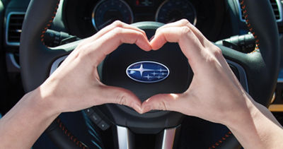 Heart shaped hands around Subaru badge on steering wheel.
