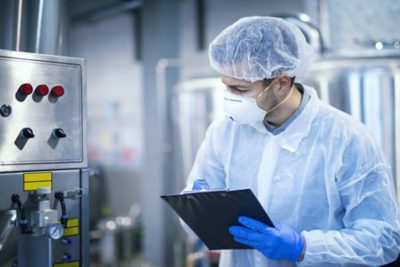 Technologist expert in protective uniform with hairnet and mask taking parameters from industrial machine in food production plant.