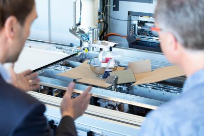 two workers watching item be packaged by machine