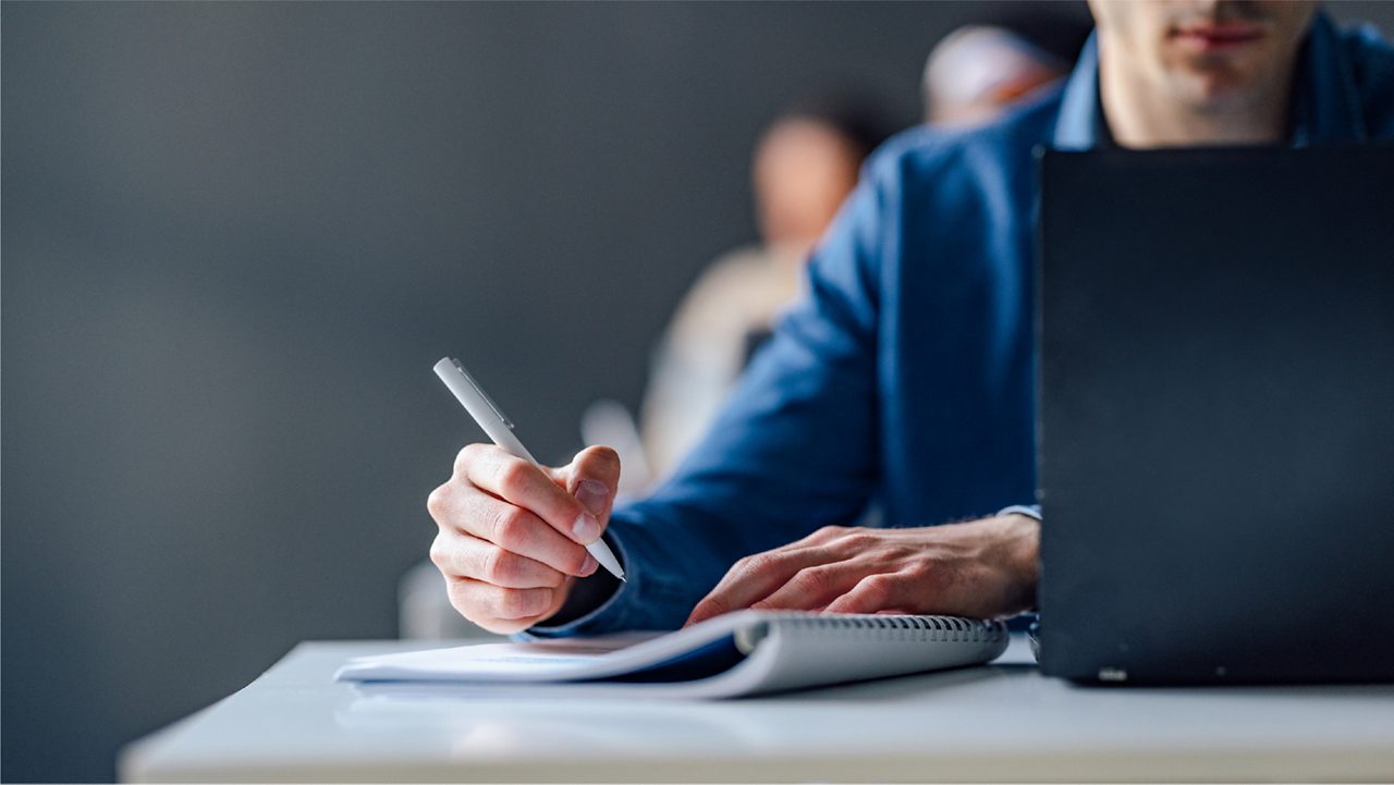 man writing on a notepad next to a laptop