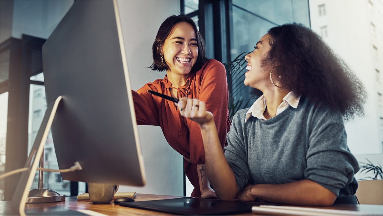 two women smile while speaking in front of a computer