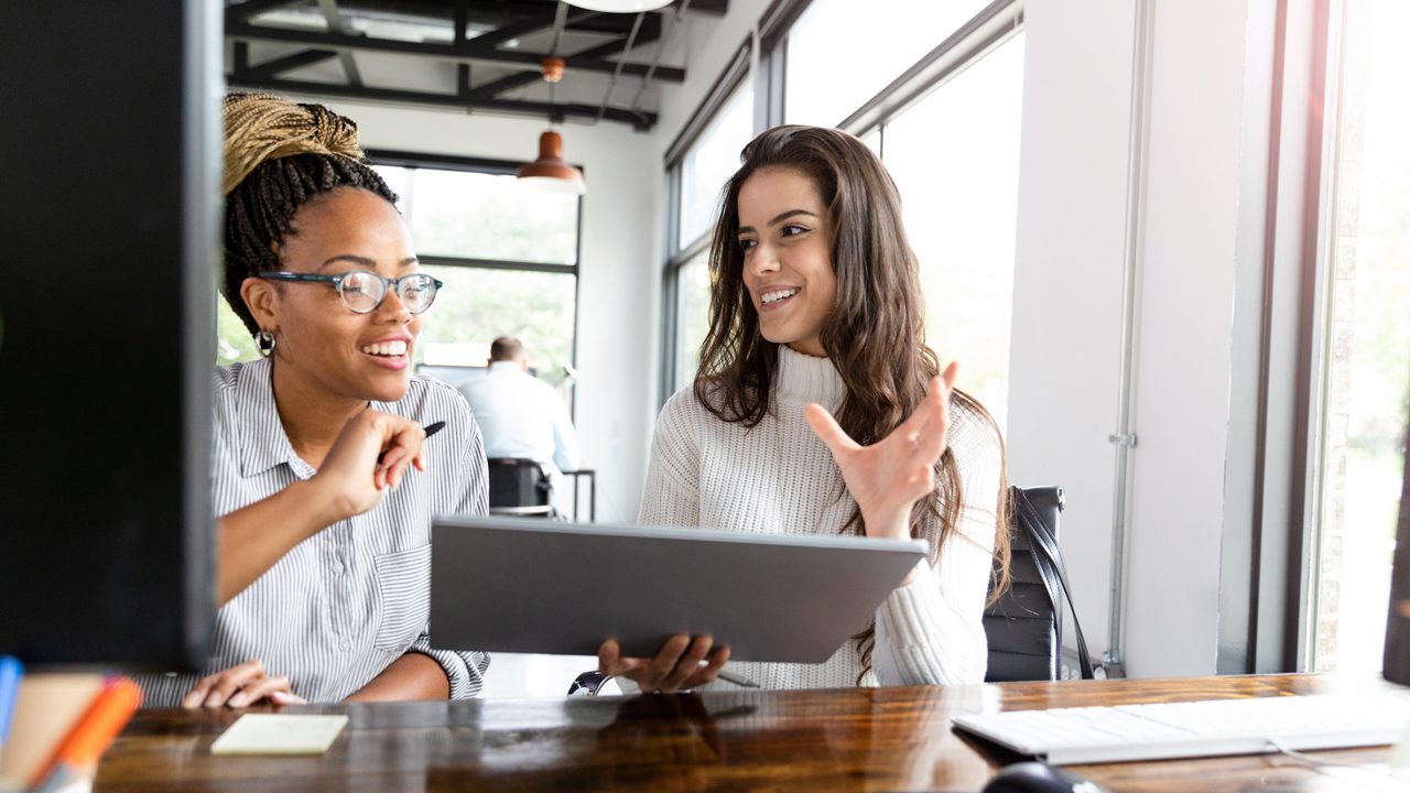 two women in an office speaking while looking at a tablet computer