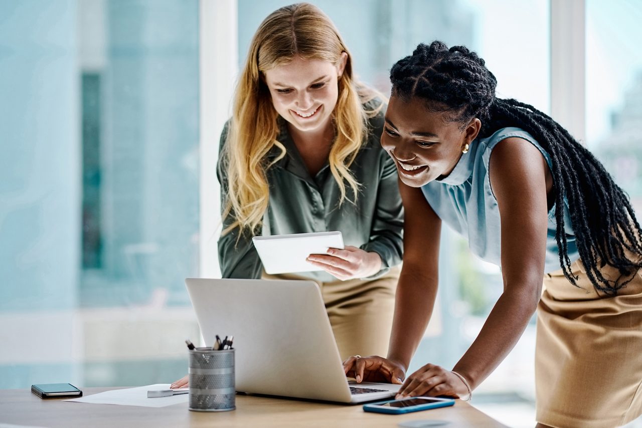 two women smile while working on a computer together in a modern office