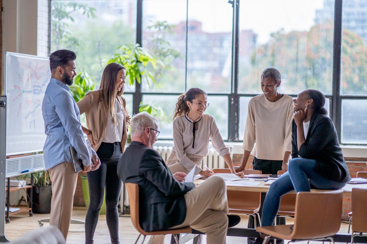 A small group of business professionals casually meet in a downtown office as they discuss new strategies and goals for the company.  They are each dressed professionally and are casually sitting around and on a boardroom table.