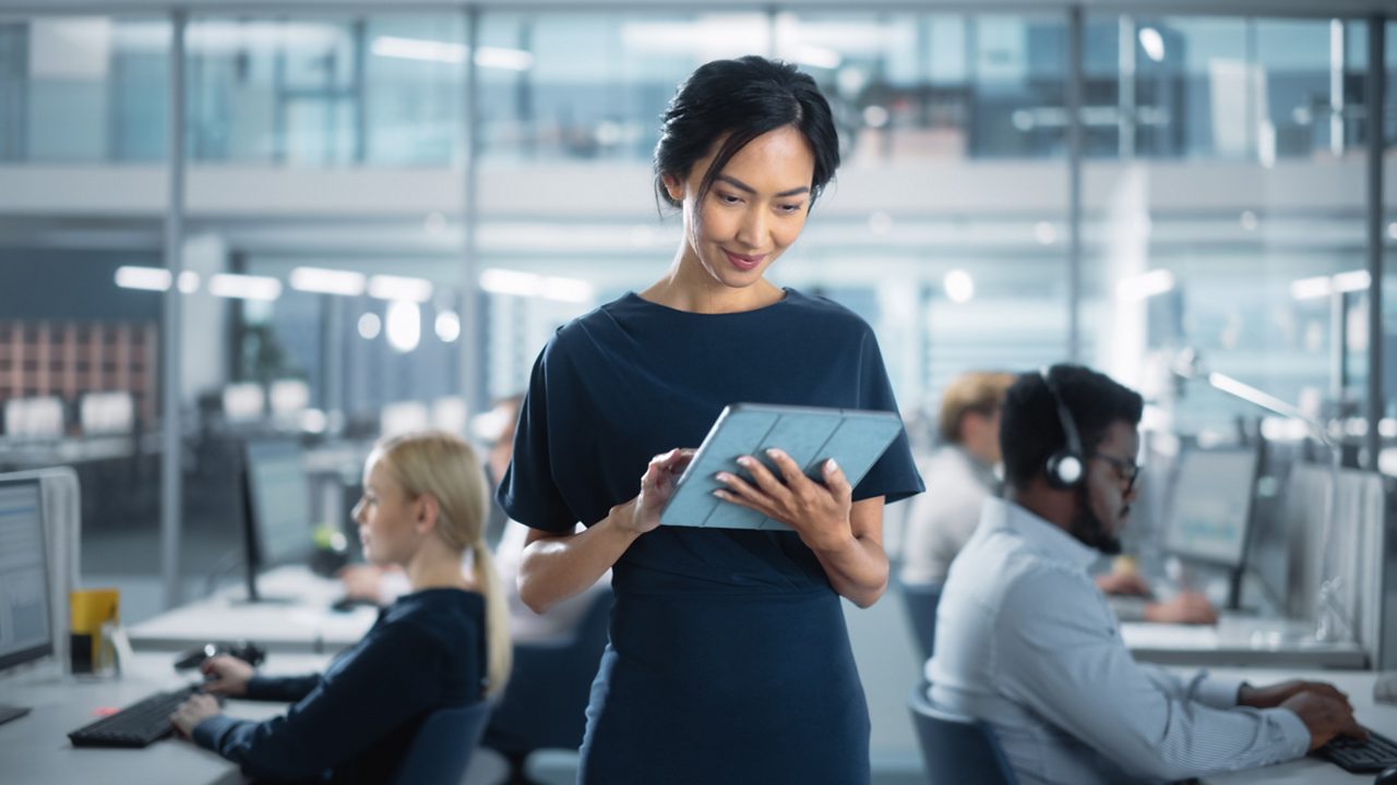 businesswoman holding tablet in front of coworkers working at their desks