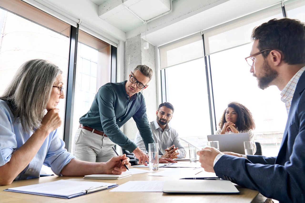 Male mature caucasian ceo businessman leader with diverse coworkers team, executive managers group at meeting. Multicultural professional businesspeople working together on research plan in boardroom.