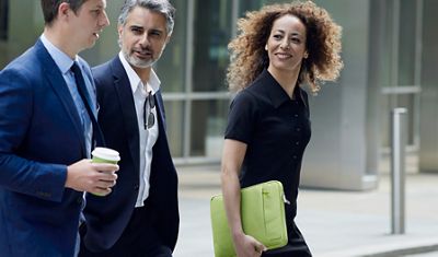 Three business professionals walking together outside a modern office building, with a woman smiling and carrying a green laptop sleeve.