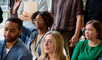 A diverse group of people seated at a community or workplace gathering, with a woman raising her hand to participate in a discussion.