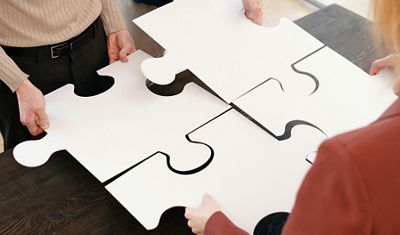Three people collaboratively fitting together large white puzzle pieces on a wooden table, symbolizing teamwork and problem-solving.