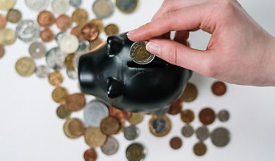 A hand inserting a coin into a black piggy bank surrounded by scattered coins of various denominations, representing savings and financial planning.