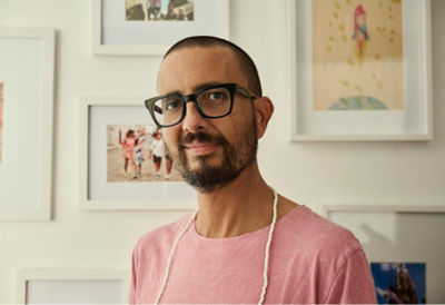 A man wearing glasses and a pink shirt stands in front of a wall adorned with various pictures.