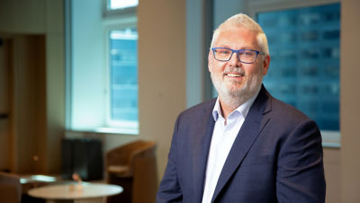 Smiling man with gray hair and blue glasses wearing a dark suit in an office setting