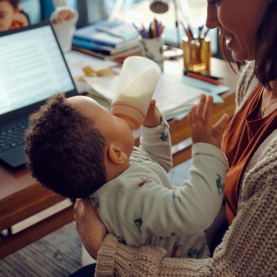 A young mother sitting at a desk with a computer, holding and feeding her infant a bottle of milk.