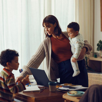A young Mom holding an infant, while helping you school-aged boy at the dining room table.