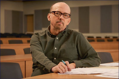 Man wearing glasses sitting in a lecture hall with papers on the table and a pen in hand