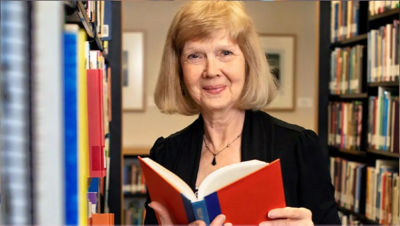An older woman standing in a library holding a red book open and smiling