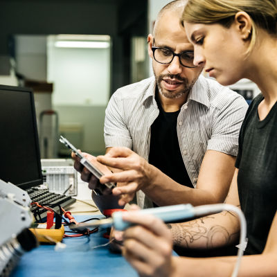 A man in a button down shirt giving a young lady direction while she holds a soldering iron.