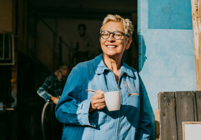 A woman wearing glasses and a denim jacket holds a coffee cup, smiling while standing outdoors.