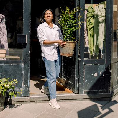 A woman stands outside a store, holding a potted plant and smiling at the camera. 