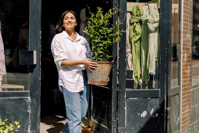 A woman stands outside a store, holding a potted plant and smiling at the camera.