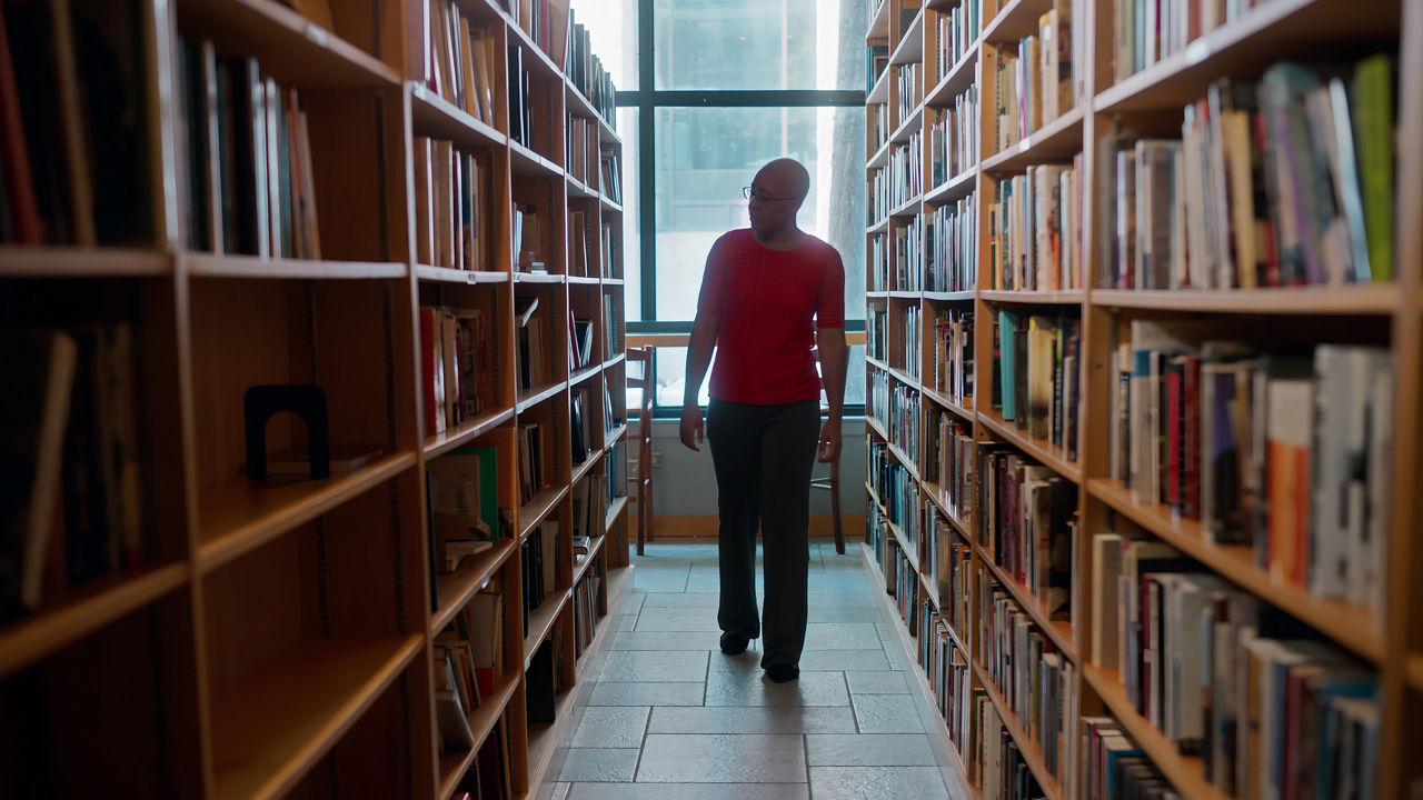 A short-haired woman with glasses walking down an aisle of bookshelves