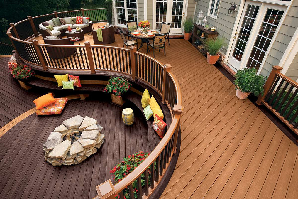 A spacious, curvilinear wooden deck featuring a lounging area, dining set, potted plants, and a stone fire pit.
