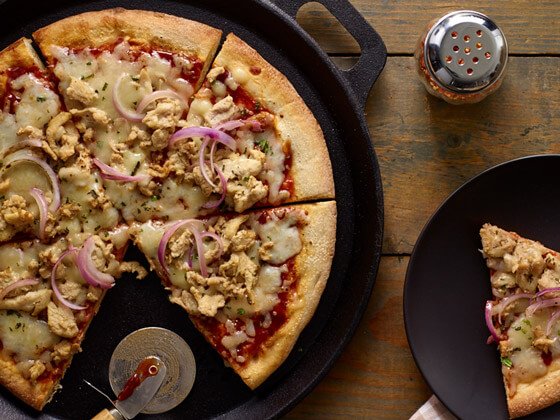 Pizza with sauce, cheese, chicken, red onion on a cast iron and red pepper flakes on the side 