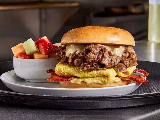 Plated beef steak, egg, bacon and cheese bagel sandwich with a side of fruit