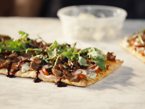 Steak-EZE® Redi Steak® Fully Cooked Seasoned Sliced Beef on top of a cherry tomato sauce topped with slices of mozzarella cheese and chunks of Bleu cheese, fresh shaved red onions, baby arugula, and drizzled with balsamic vinegar and olive oil all served on a focaccia style flatbread crust.