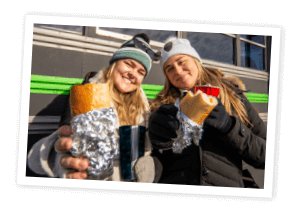 Two girls with jackets and hats, holding a philly sandwich 