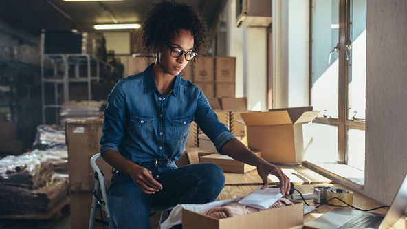 Female sitting on a stool and is preparing an order surrounded by shipping supplies 