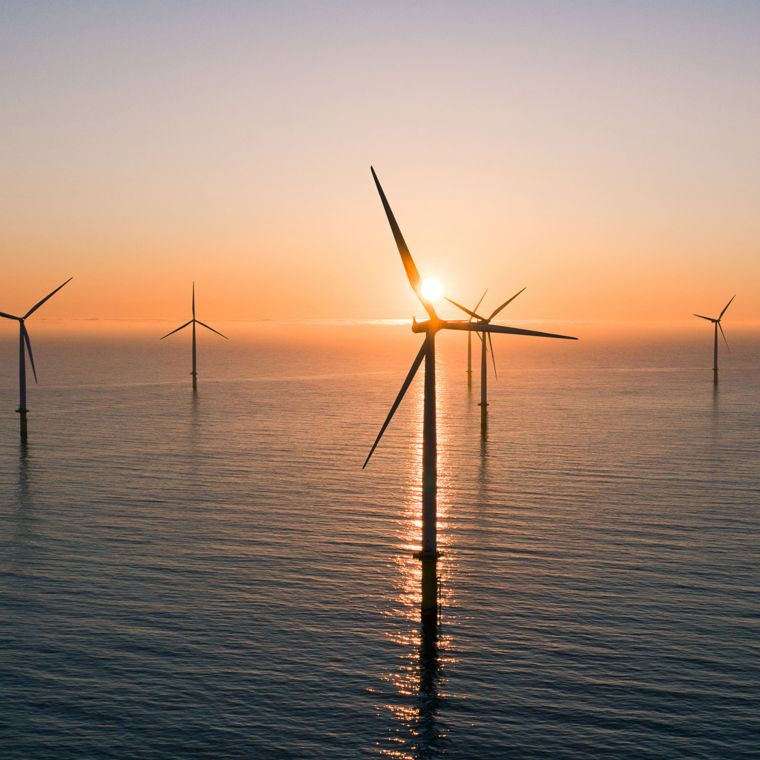 A group of wind turbines emerge from the ocean surface as the sun sets behind them.
