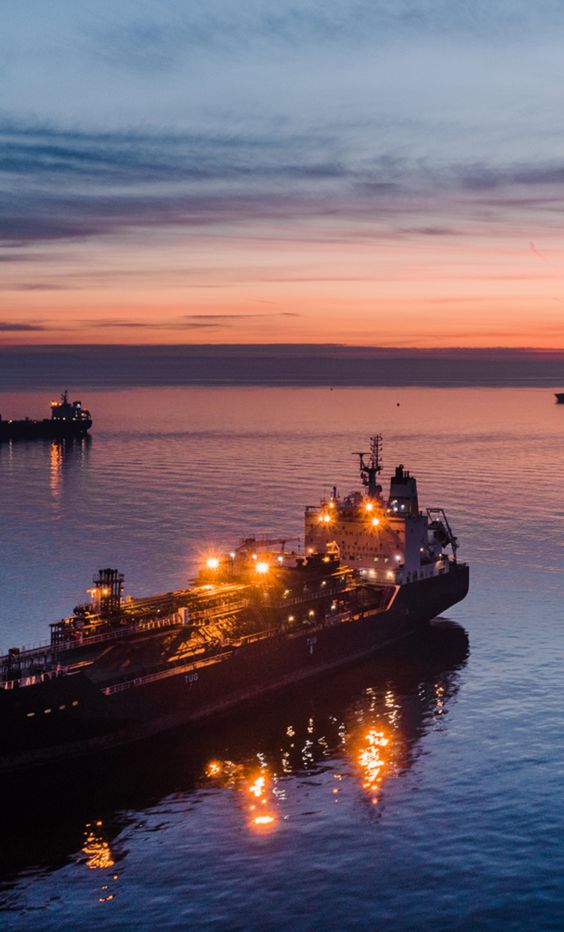 As evening falls, an oil tanker moves through the ocean, with other ships nearby.