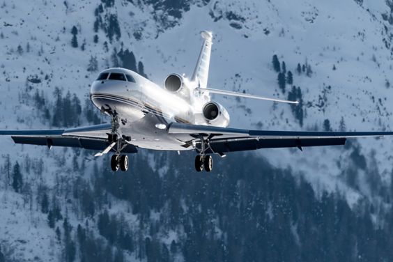 A three-engine private jet in flight with its wheels down, with a snowy, forested mountain behind it.
