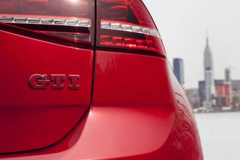A red Volkswagen Golf GTI is shown from the back, with badge in closeup against a city skyline.