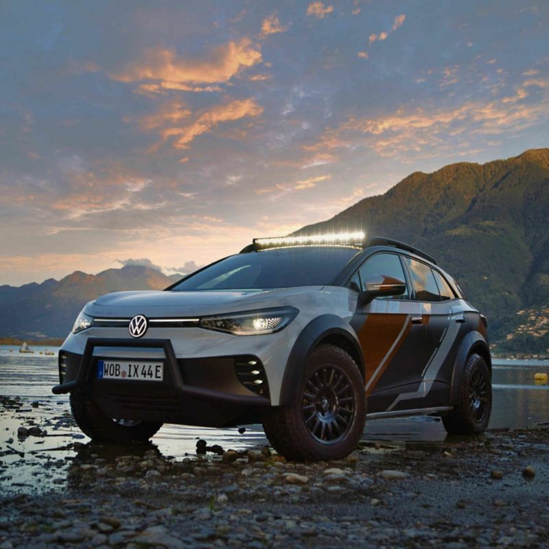 An angled shot of the ID. XTREME concept car in metallic white and strips of metallic red and blue situated on a pebble-lined beach with mountains and a rolling purple, blue sky in the background. 