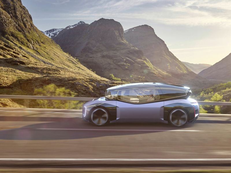 A futuristic looking concept car called the GEN.TRAVEL with a distinct design divided into two parts: the top features a transparent, glass cabin and the body, a metallic purple. The car drives down a scenic mountain highway. 