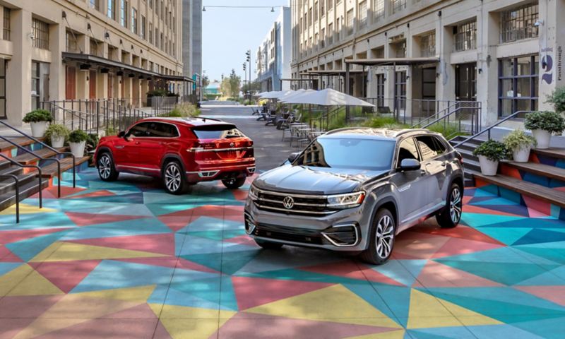 Two Atlas Cross Sports, one in Aurora Red Metallic and one in Pure Gray, parked in a colorful courtyard in between two tall buildings.