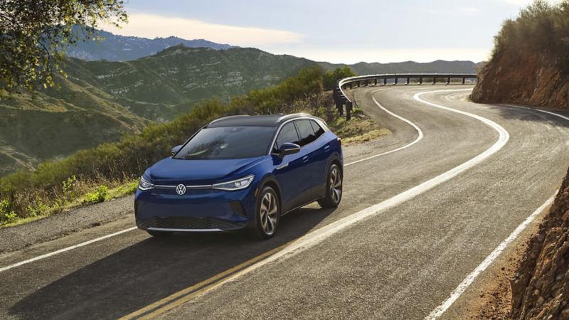 A dusk blue metallic Volkswagen ID.4 curves along an open road surrounded by a verdant, hilly landscape. 