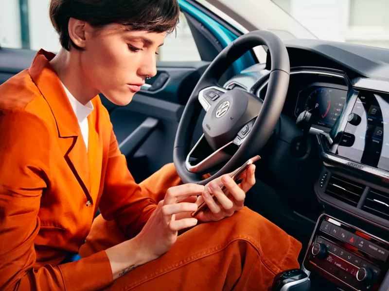 A chic young woman in a casual burnt orange pantsuit sits in the driver’s seat of a parked vehicle scrolling through phone.