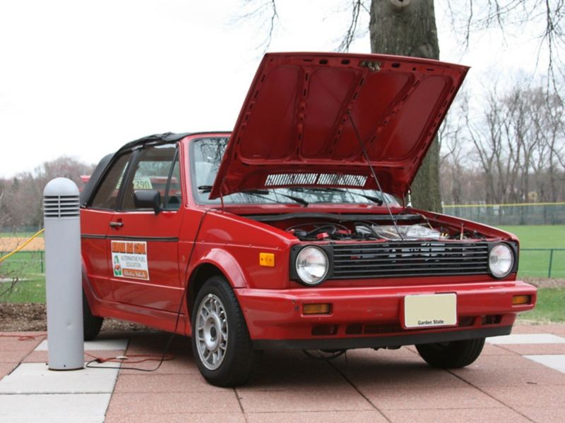 A red 1990 Volkswagen Cabriolet that has been converted from gas to electric is parked with its hood open.