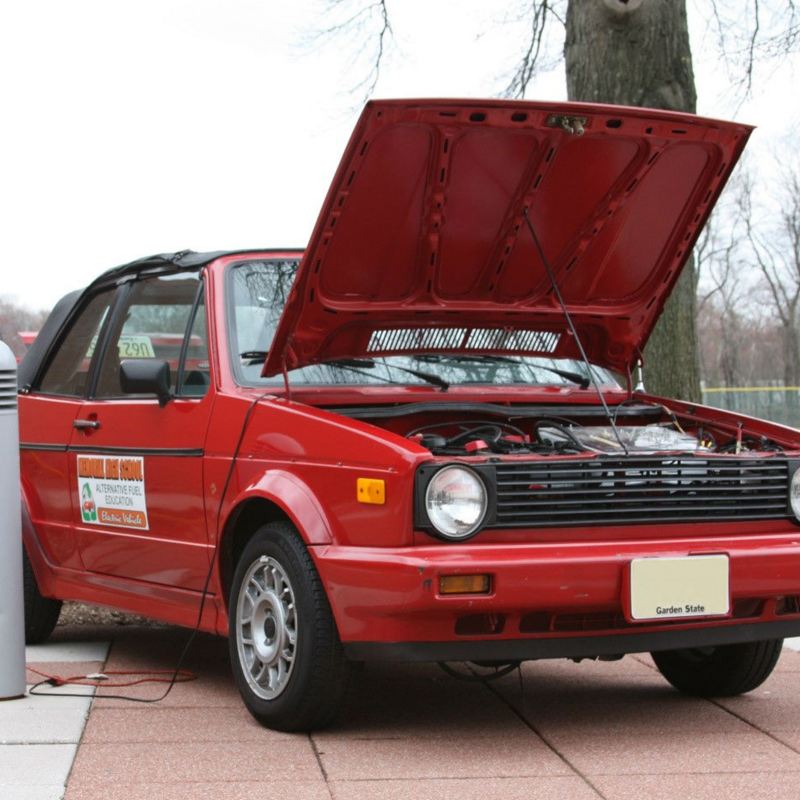 A red 1990 Volkswagen Cabriolet that has been converted from gas to electric is parked with its hood open.